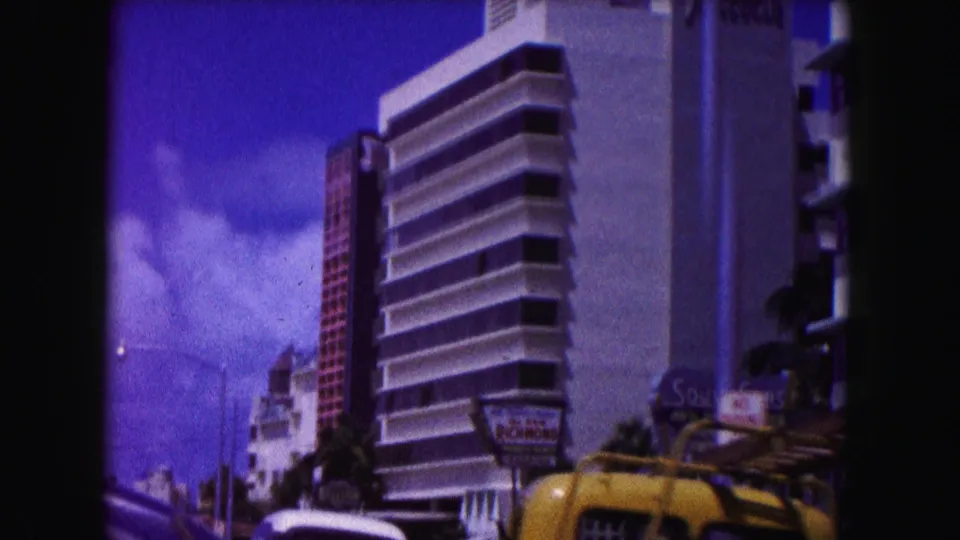 1960 Miami street view with apartment towers, a yellow taxi, hotel signage, and bright blue sky captured on 8mm film.