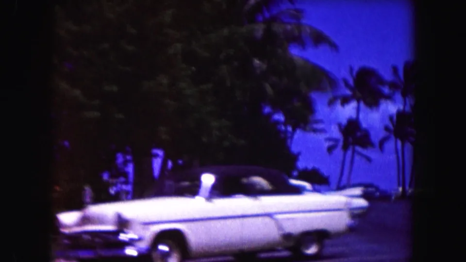 1960 Miami street frame with a white classic convertible, palm silhouettes, dark foliage, and saturated blue sky.