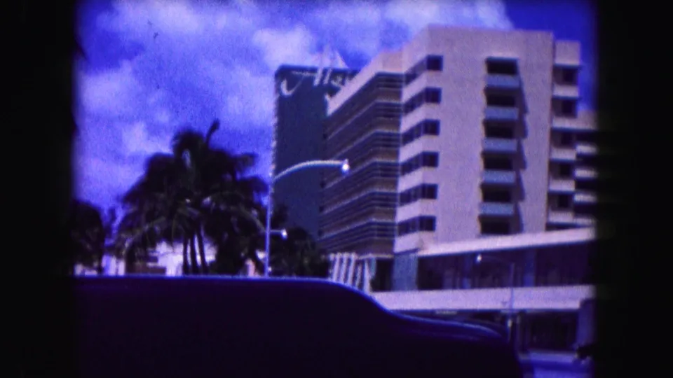 1960 Miami street view with palm trees, hotel balconies, and a dark car edge crossing the foreground.