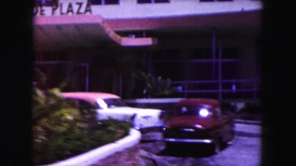 1960 Miami street-level hotel entrance with partial plaza signage, parked cars, curved curb, and tropical landscaping.