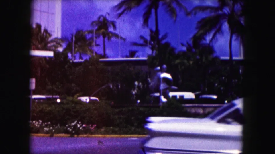 1960 Miami home-movie frame with a classic car near palms, hedges, and a modern building facade.
