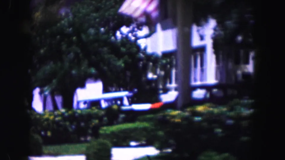 1960 Miami home-movie frame showing a residential facade, parked cars, palms, and shrubs from a moving car.