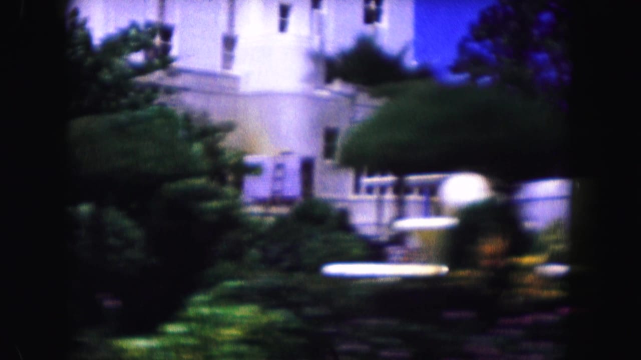 Pale building partly obscured by trees and landscaping in soft, bluish 8mm footage.