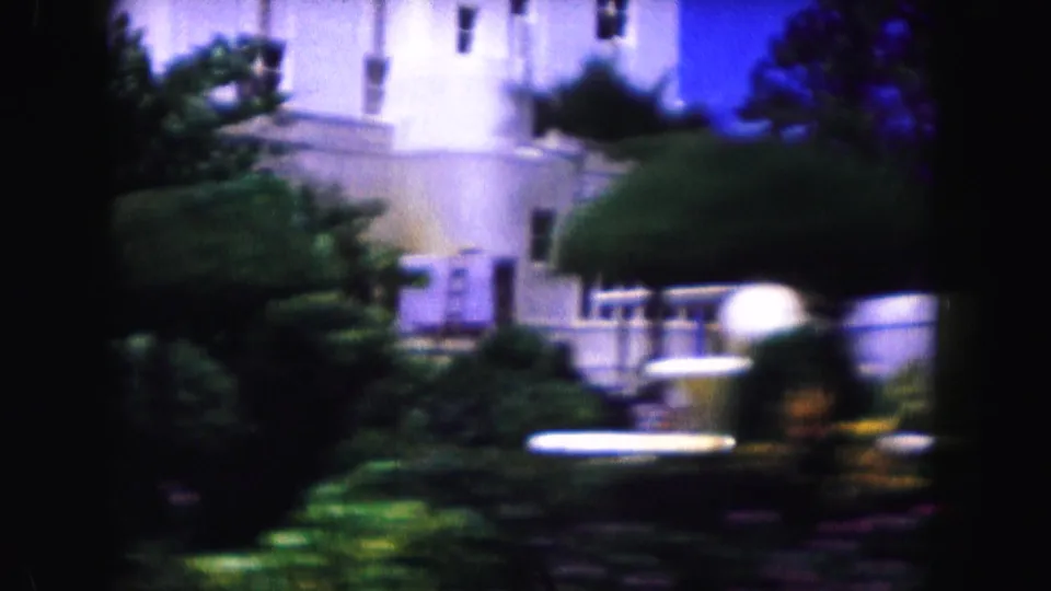 1960 Miami home-movie view of a pale building behind dense trees and trimmed landscaping, filmed with soft focus and a blue-purple cast.