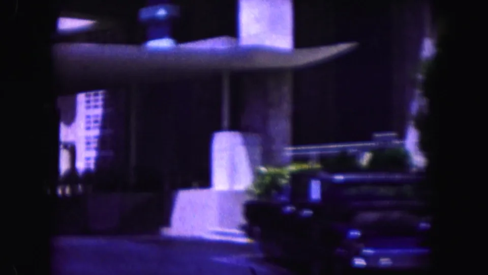 1960 Miami home-movie frame of a modern-looking exterior with columns, an overhang, plants, and a dark vintage car near the street.