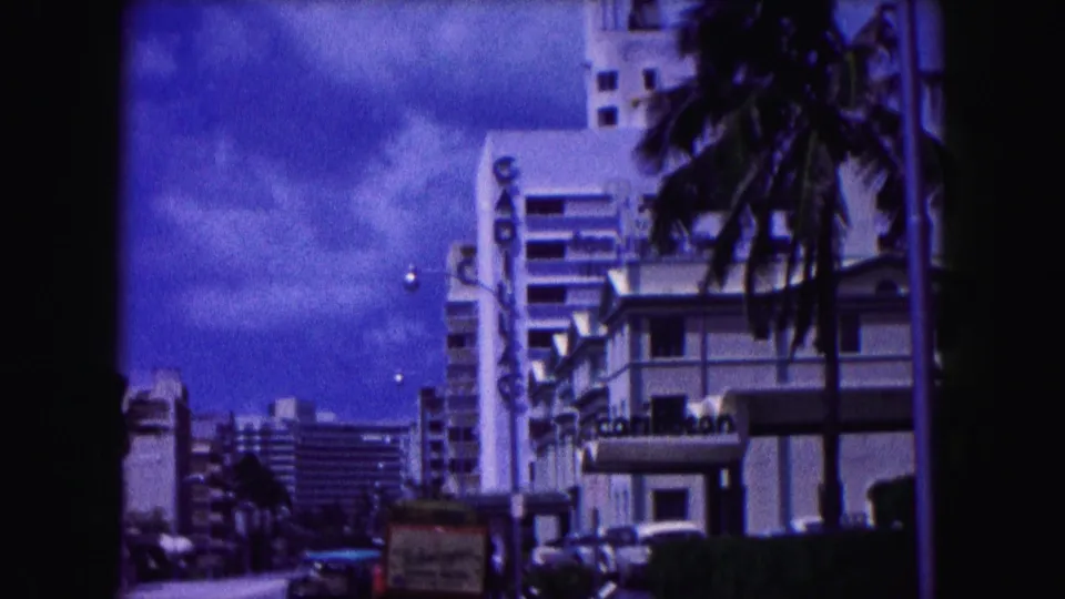 1960 Miami 8mm street scene with hotel architecture, palms, parked cars, and a small curbside sign visible in the foreground.
