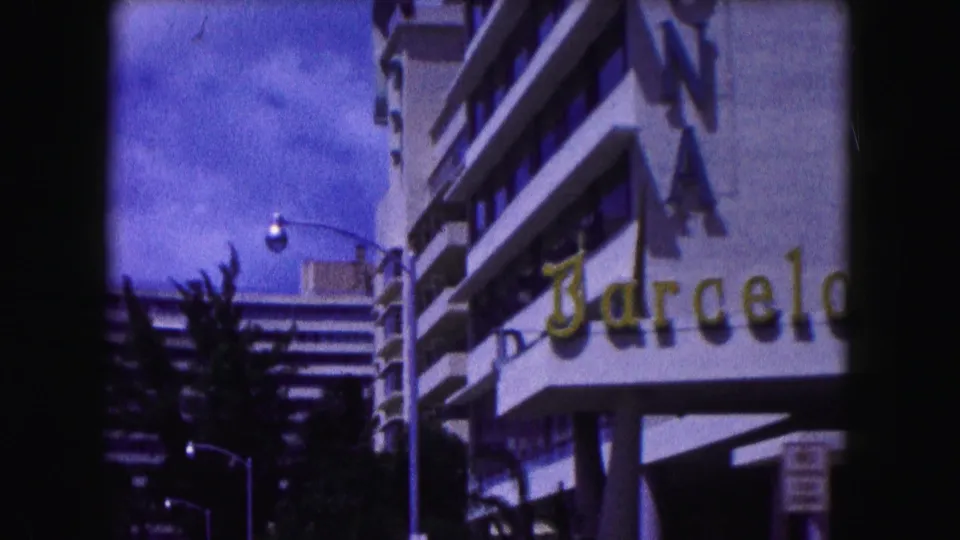 1960 Miami home-movie view of a white modernist building with a partial Barcelona sign, palms, streetlamps, and bright sky.