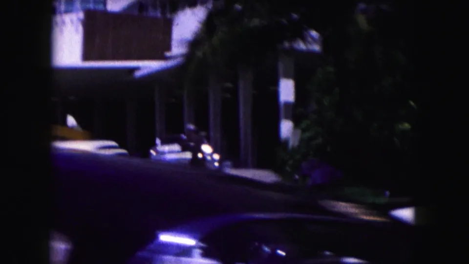 1960 Miami home-movie frame with parked American cars, palm foliage, a shaded sidewalk, and a white building entrance area.