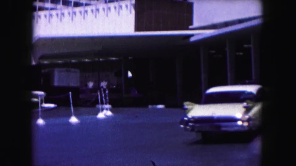 1960 Miami street scene with a white building frontage, parked mid-century car, street signs, and a pedestrian near the sidewalk.
