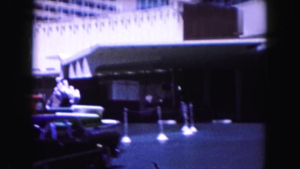 1960 Miami drive-by frame with classic cars, a building facade, sidewalk posts, and small pedestrians or figures in soft 8mm color.