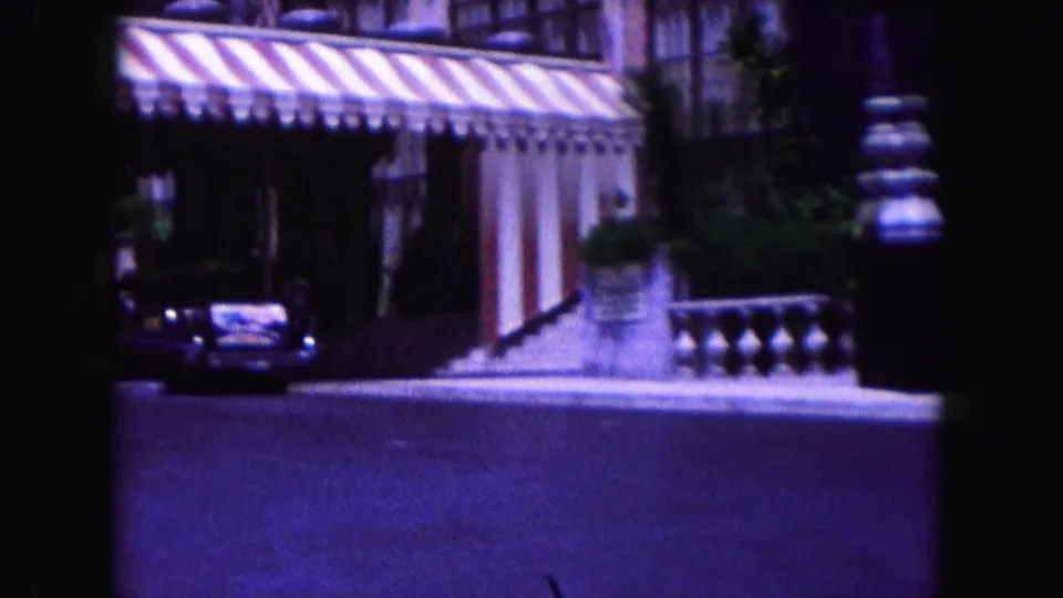 1960 Miami home-movie frame showing a striped awning, building facade, balustrade, greenery, and a white car near the curb.