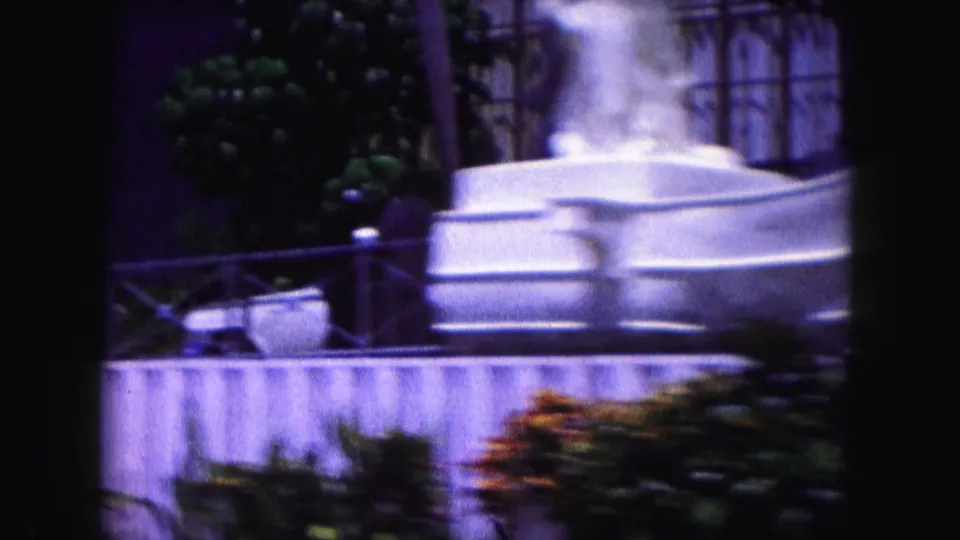 1960 Miami home-movie frame showing a pale decorative fountain, low wall, plants, and windows in soft 8mm focus.