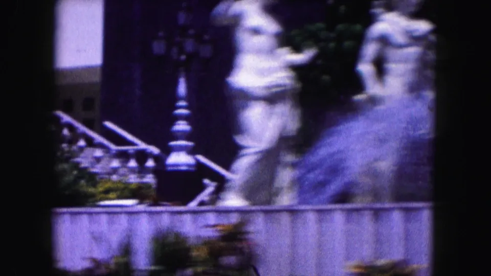 1960 Miami exterior frame with two pale garden statues, a low wall, greenery, and dark architectural posts in a home-movie scan.