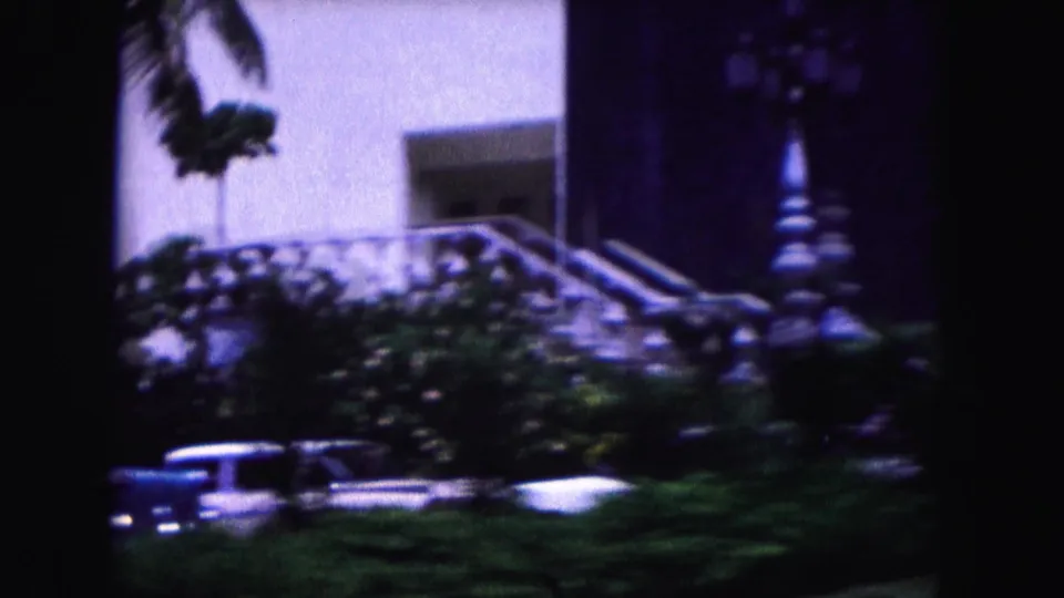 1960 Miami 8mm frame with palm foliage, a white exterior stair area, and mid-century cars blurred by a passing camera.