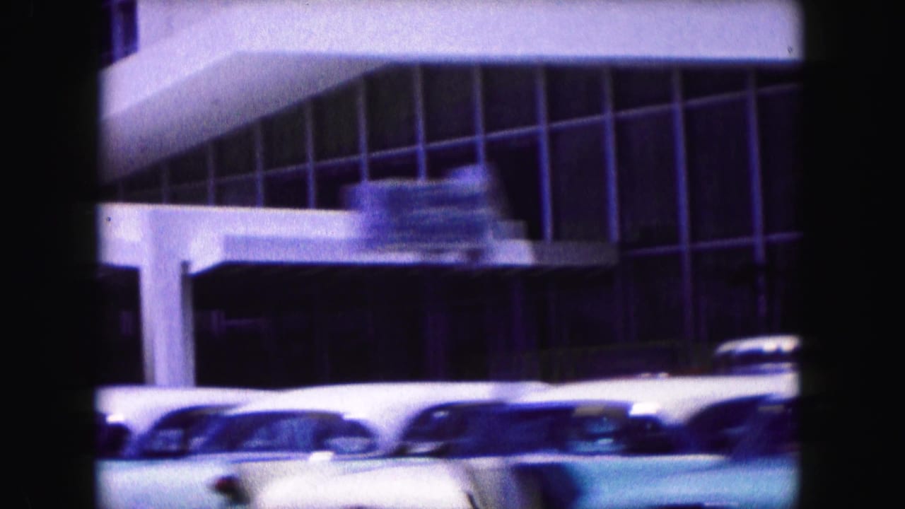 Modernist glass-fronted building with parked mid-century cars and a blurred vehicle in front.