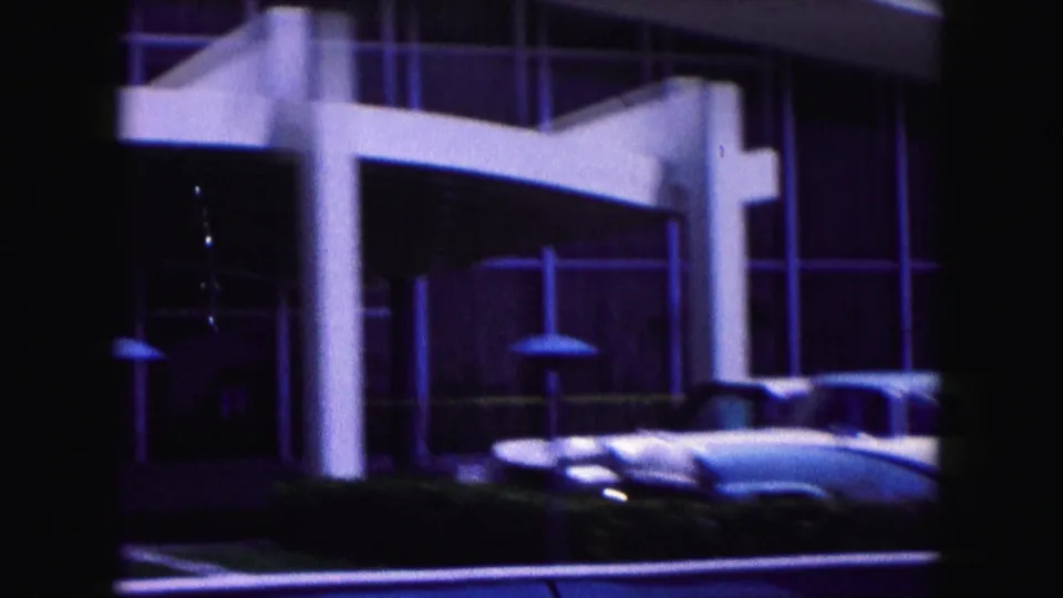 1960 Miami exterior with a modernist entrance, white columns, large glass panes, and parked classic cars along the curb.