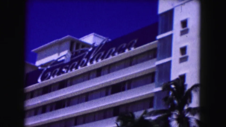 1960 Miami home-movie frame showing a white hotel facade, dark palm shapes, and a deep blue sky from a passing street view.