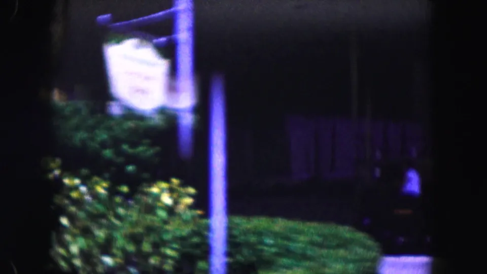 1960 Miami home-movie frame with a bright roadside hotel sign, landscaping, lawn, and shadowed entrance area seen from motion.