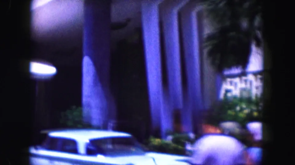 1960 Miami hotel exterior with entrance columns, a parked white car, palms, and a partially visible blurred person at the lower right edge.