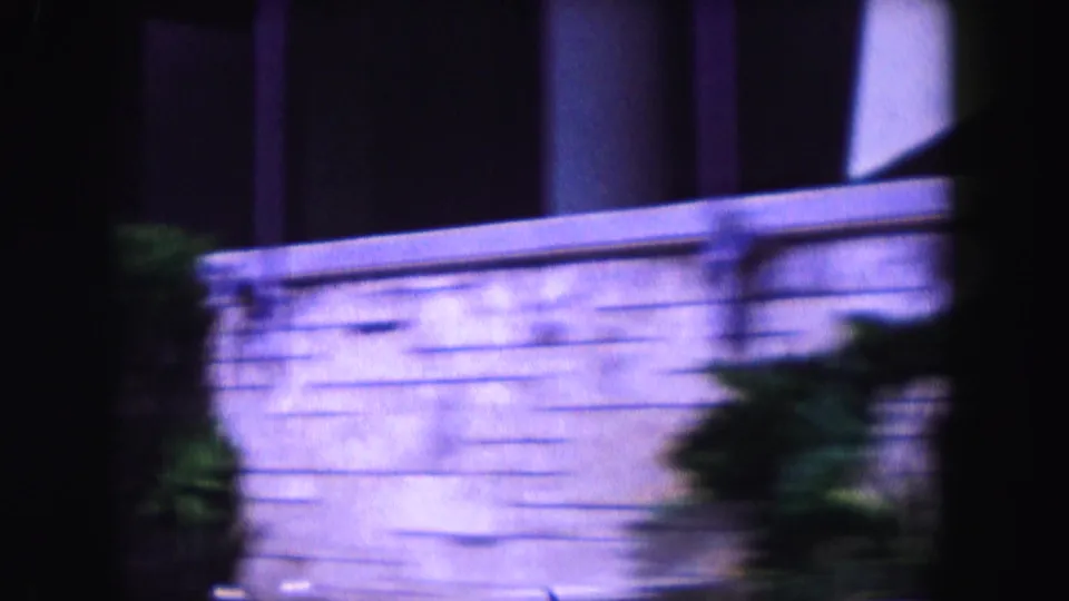 1960 Miami 8mm frame showing a wall or building edge with foliage, dark vertical foreground shapes, and heavy purple-blue color shift.