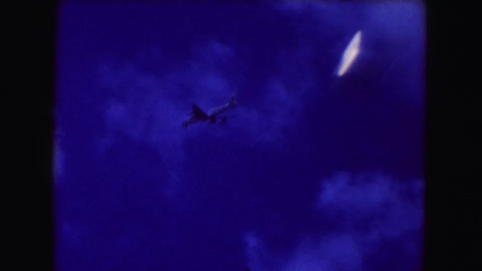 1960 Miami 8mm frame with a dark airplane silhouette against a grainy blue sky and soft cloud patches.
