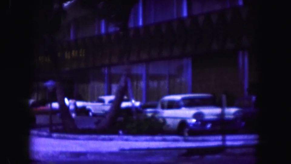 1960 Miami street frame showing parked vintage cars along a low building, partly obscured by dark foreground shapes and blue-purple film color.
