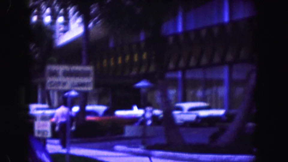 1960 Miami-area 8mm frame showing classic cars, a hotel facade, sidewalk signage, and a person near the curb.