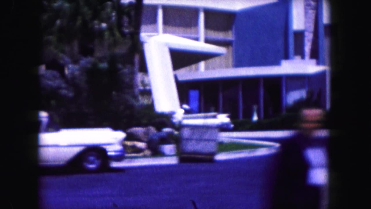 Modernist hotel entrance with a white car, palm landscaping, and a blurred passerby near the street.