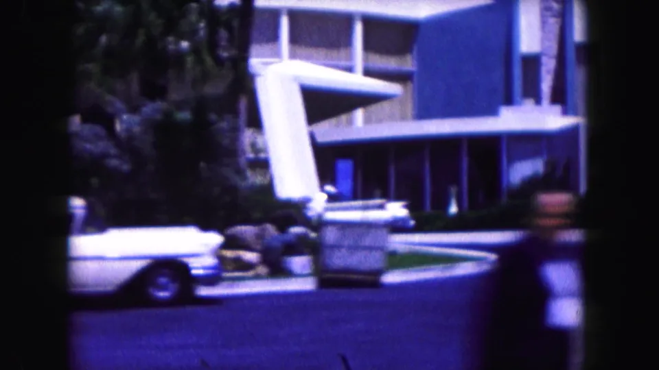 1960 Miami hotel entrance with angular facade, white classic car, palm landscaping, and handheld home-movie blur near the foreground.