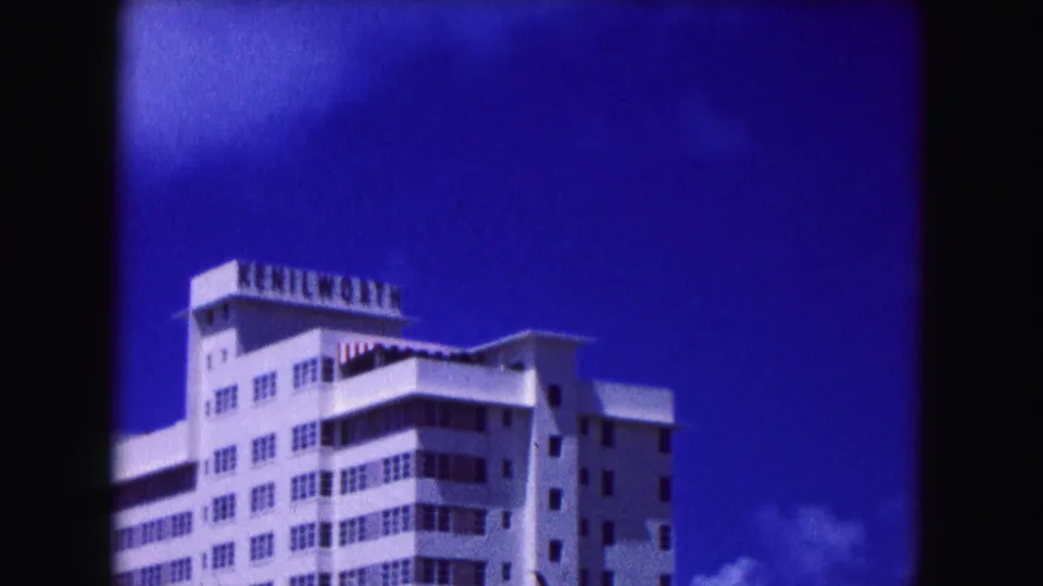 1960 Miami hotel tower with rooftop sign, balconies, rows of windows, and saturated blue sky in 8mm color.