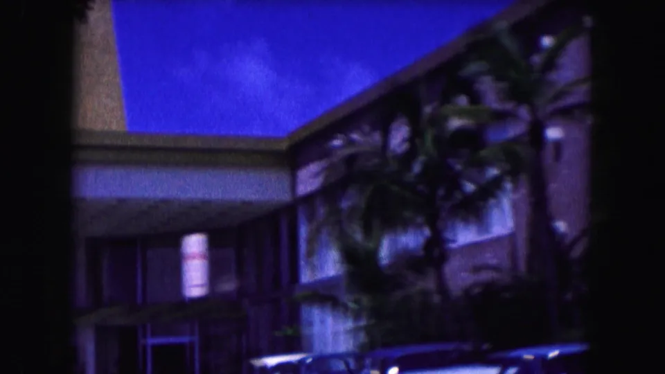 Miami hotel exterior in 1960 partly hidden by palms, with parked classic cars, windows, roofline, and handheld 8mm softness.