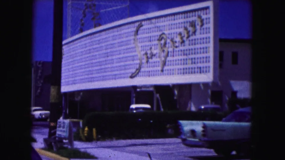 1960 Miami home-movie frame showing a large commercial sign, curbside cars, and dark vertical film-gate borders.