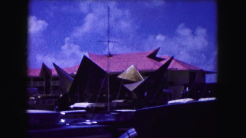 1960 Miami street view showing a mid-century building with angular roof elements, parked cars, and a bright clouded sky.