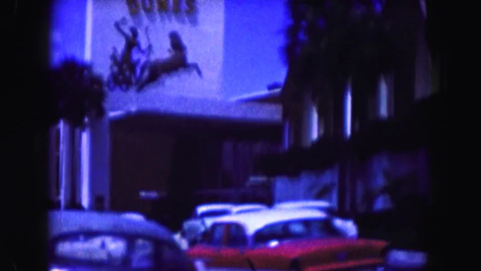 1960 Miami roadside commercial exterior with parked mid-century cars, a red car, and a low building under strong blue color shift.
