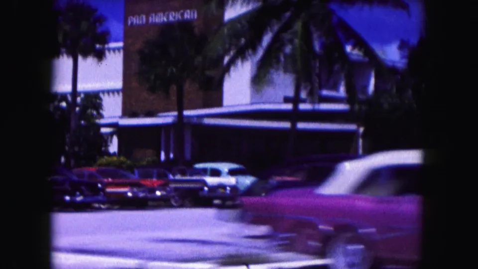 1960 Miami street scene showing palm trees, parked vintage cars, and a pink convertible crossing the frame.