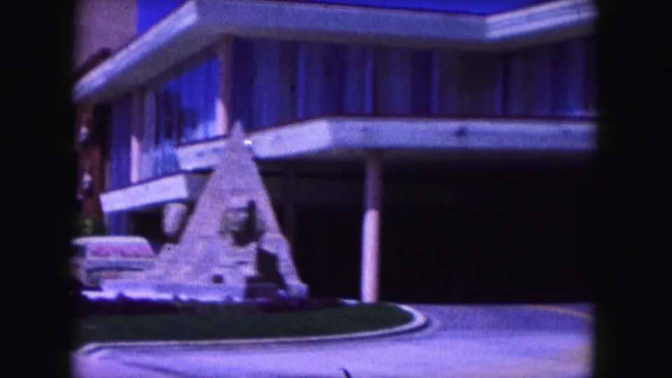 1960 Miami 8mm frame with a themed roadside sculpture, modern building exterior, roadway, and partial vintage car.
