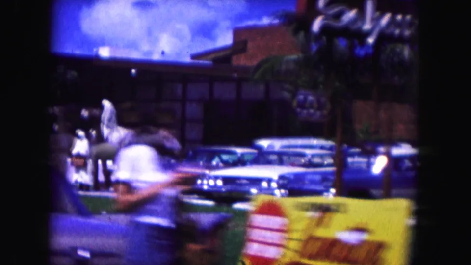 1960 Miami frame showing a sphinx-like roadside sculpture, classic cars, signage, and a low building.