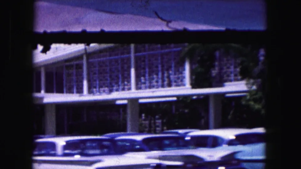 1960 Miami home-movie frame with parked classic cars below a modernist facade, partly obscured by dark film borders.