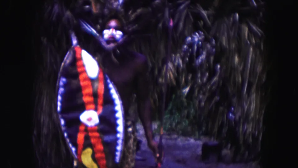 1960 Miami 8mm frame showing a costumed figure with a decorated shield and spear-like prop standing near dense foliage.