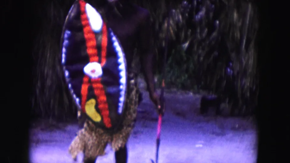 1960 Miami 8mm frame showing a costumed person with a feathered headdress, staff, and red, white, and yellow shield near palms.