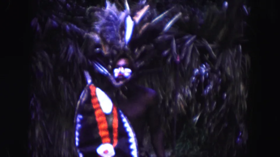 1960 Miami 8mm frame showing a closer visitor view of a costumed person with shield, staff, feathered headdress, and palm leaves.