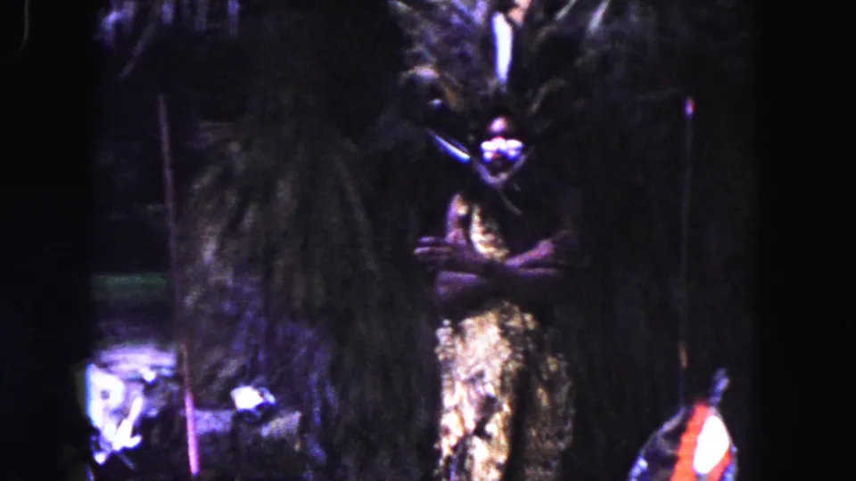 1960 Miami home-movie frame showing a costumed figure beside thatch and posts, with the face area partly obscured by blur and shadow.