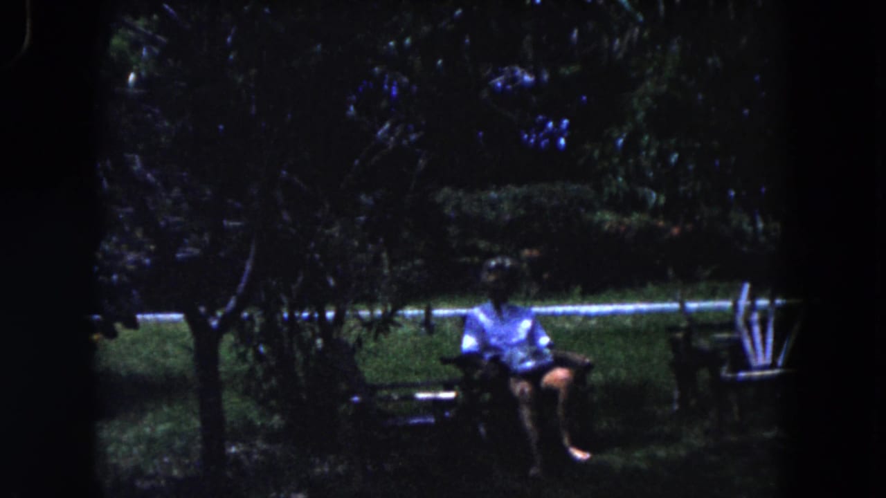 Man seated on a bench in a dark garden with trees and lawn around him.