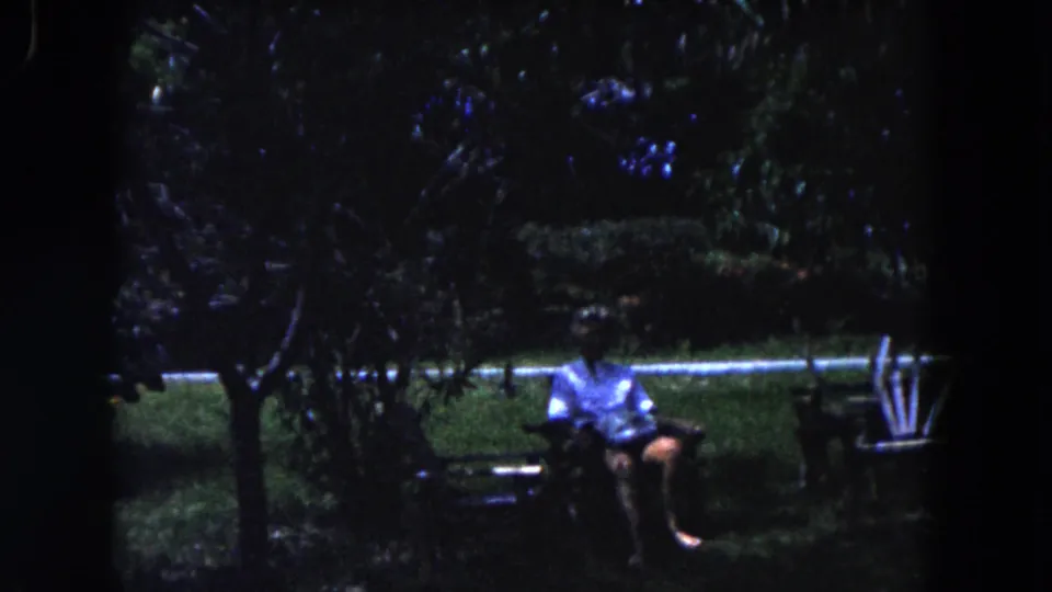 1960 Miami home-movie frame showing a man in light clothing seated on a garden bench beneath trees.