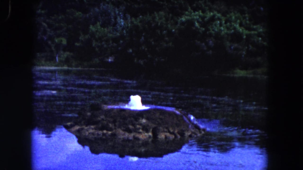 Small water spray emerging from a rock mound in a lake with trees behind it.