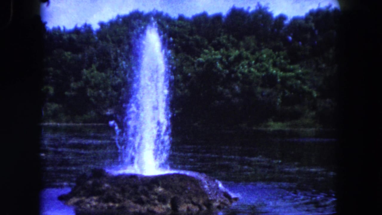 Tall water jet rising from a rock mound in a lake with trees in the background.