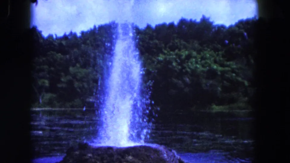 1960 Miami-area 8mm frame with a bright vertical water column spraying from a rock in the lake, with trees and film grain visible.