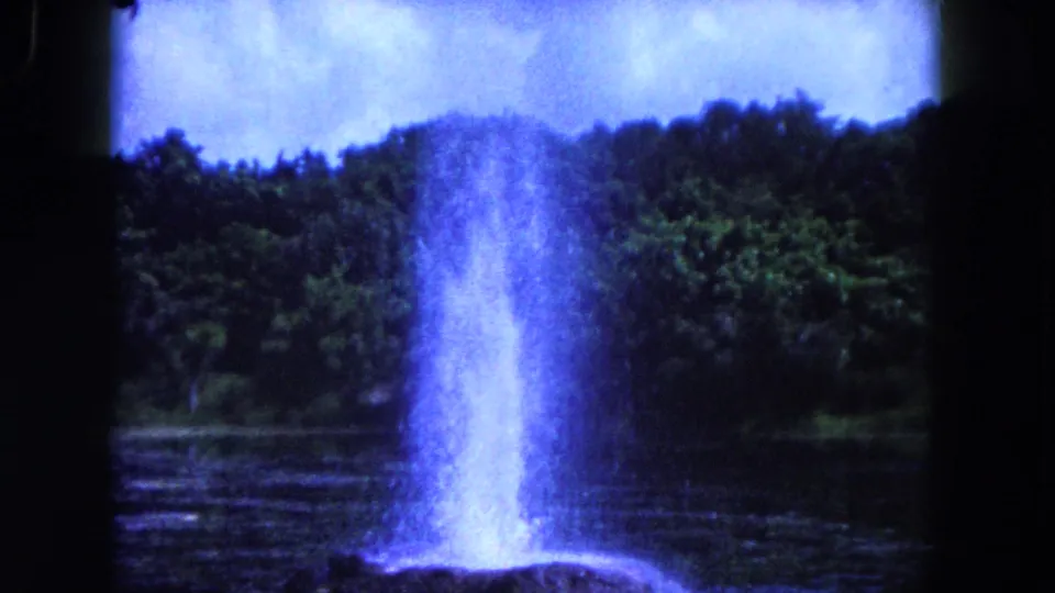 1960 Miami-area 8mm frame showing the geyser-like spray widening above the rock mound, with lake water and trees behind it.