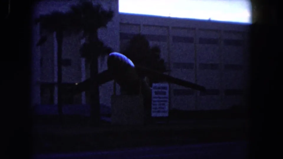 1960 Miami home-movie frame showing an indistinct silhouetted person near a roadside building and palms.