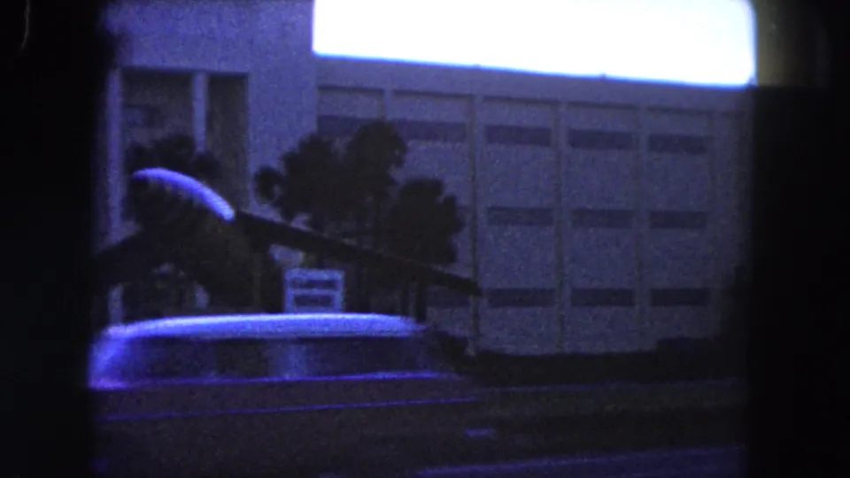 1960 Miami home-movie frame showing an aircraft display in front of a modern building, with palm trees and a car passing at the edge of the shot.